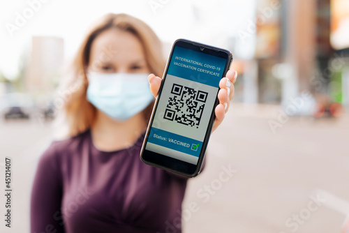 Young woman in medical face mask shows health passport on mobile phone, international covid-19 vaccination certificate close-up, outside. Selective focus on smartphone screen with qr code. New reality