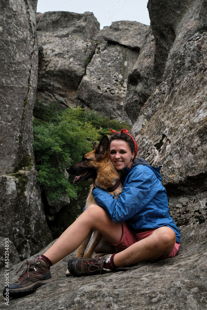 Female tourist on mountain hike in blue jacket and boots. Pretty young Caucasian woman is sitting on rock and hugging dog. Hiking in mountains with German Shepherd.