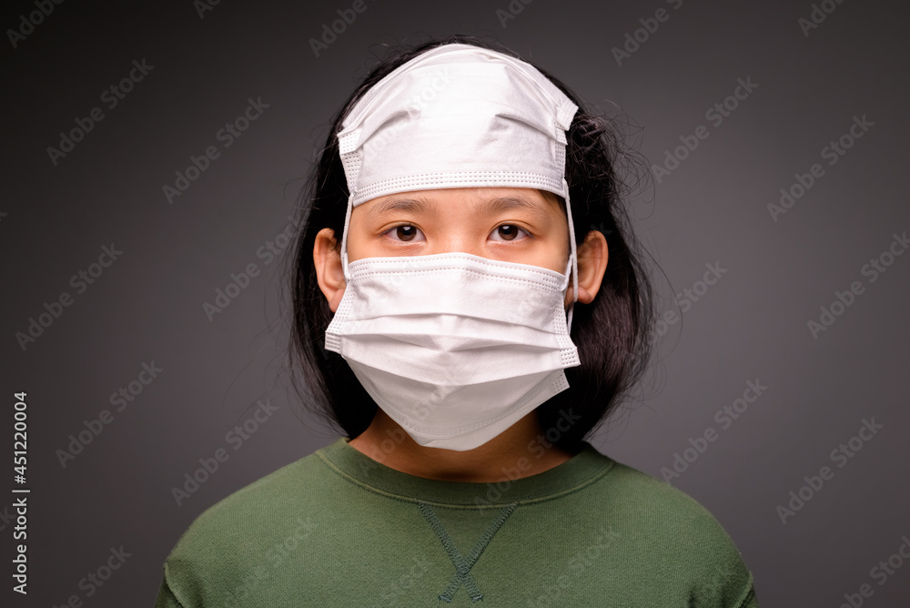 Child girl wearing two face masks for covering her nose and mouth, and another one on her head. Not over yet for covid-19 pandemic, protect the child from the waves of pandemic Child girl wearing two face masks for covering her nose and mouth, and another one on her head. Not over yet for covid-19 pandemic, protect the child from the waves of pandemic