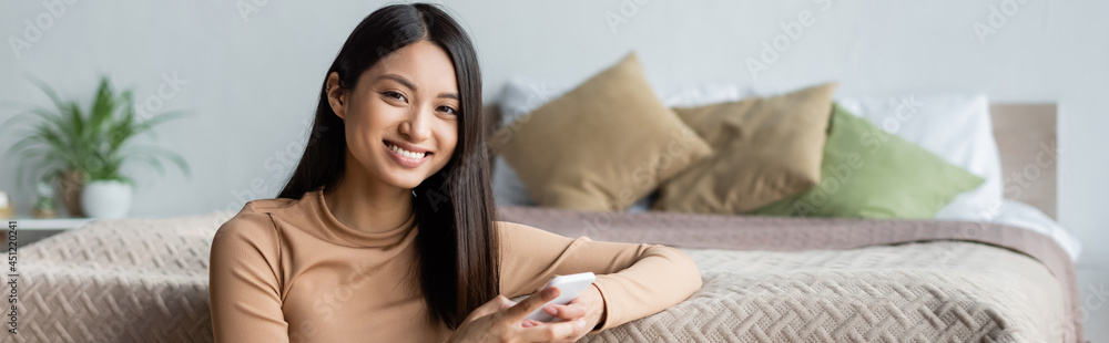 cheerful asian woman looking at camera while using smartphone in bedroom, banner. cheerful asian woman looking at camera while using smartphone in bedroom, banner.