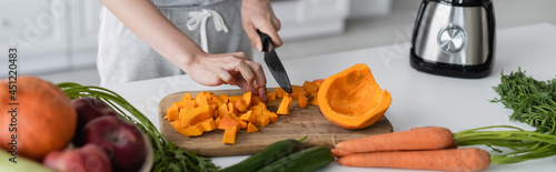 partial view of woman cutting pumpkin near fresh vegetables on kitchen table, banner.