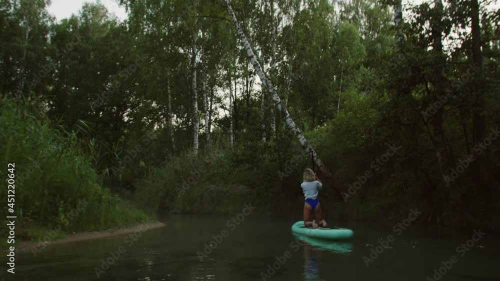Blonde woman paddling on surfing board, standing on knees, man swimming after sportswoman rowing on paddleboard, back view. Female coach rows on surfboard. Sports lesson for beginners on water. Blonde woman paddling on surfing board, standing on knees, man swimming after sportswoman rowing on paddleboard, back view. Female coach rows on surfboard. Sports lesson for beginners on water.