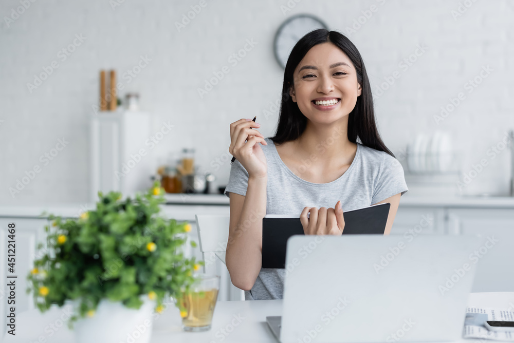 happy asian woman sitting with pen and notebook near laptop and blurred plant. happy asian woman sitting with pen and notebook near laptop and blurred plant.