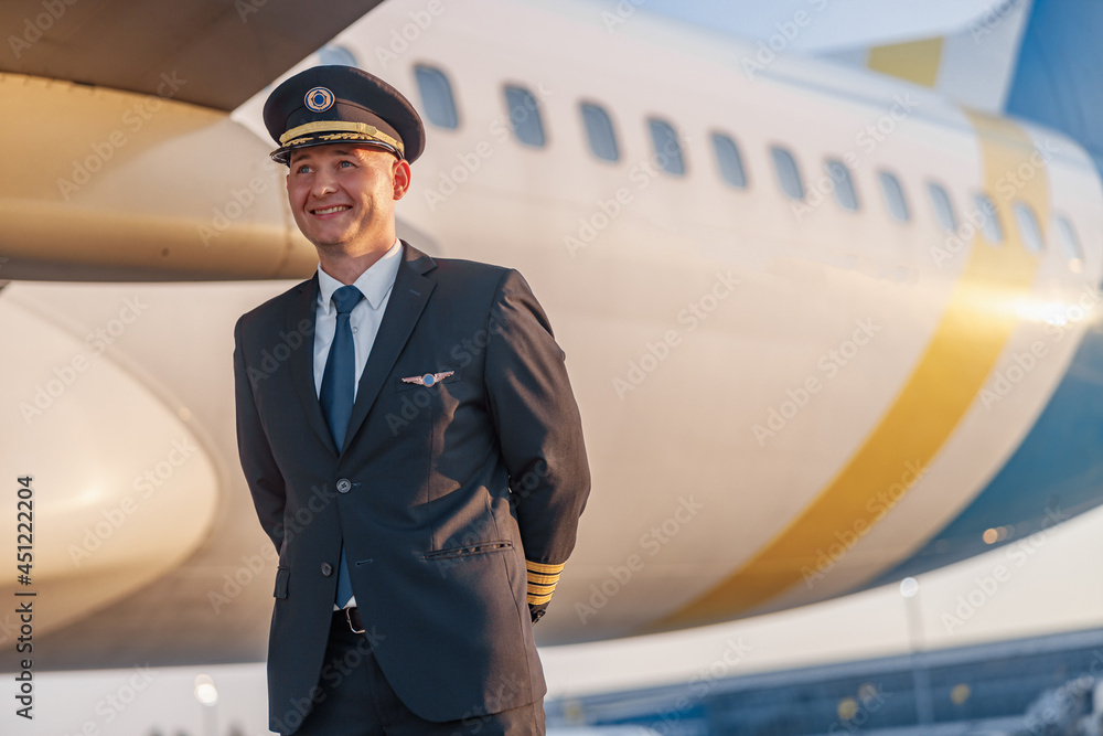 Φωτογραφία Portrait of smiling young male pilot in uniform and hat looking aside, posing in front of a big passenger airplane in airport at sunset