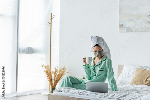 asian woman in clay mask and pajamas sitting on bed with cup of tea and looking at laptop.
