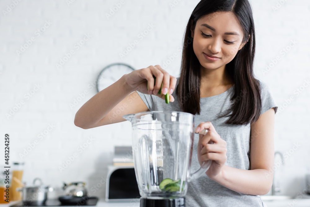 smiling asian woman adding fresh zucchini into electric shaker in kitchen. smiling asian woman adding fresh zucchini into electric shaker in kitchen.