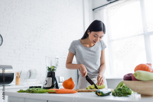 young asian woman cutting zucchini near raw vegetables and electric shaker in kitchen.