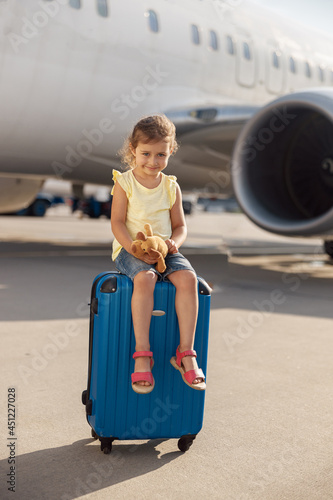 Full length shot of cute little girl holding her toy, looking at camera and sitting on suitcase in front of big airplane. Childhood, traveling, vacation concept photo