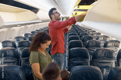 Cheerful family man putting carry on luggage in compartment while traveling together with his wife and little daughter by plane. Family vacation, transportation concept