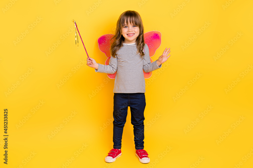 Full length photo of cute small blond hair girl hold stick wear wings shirt jeans sneakers isolated on yellow backgound Full length photo of cute small blond hair girl hold stick wear wings shirt jeans sneakers isolated on yellow backgound