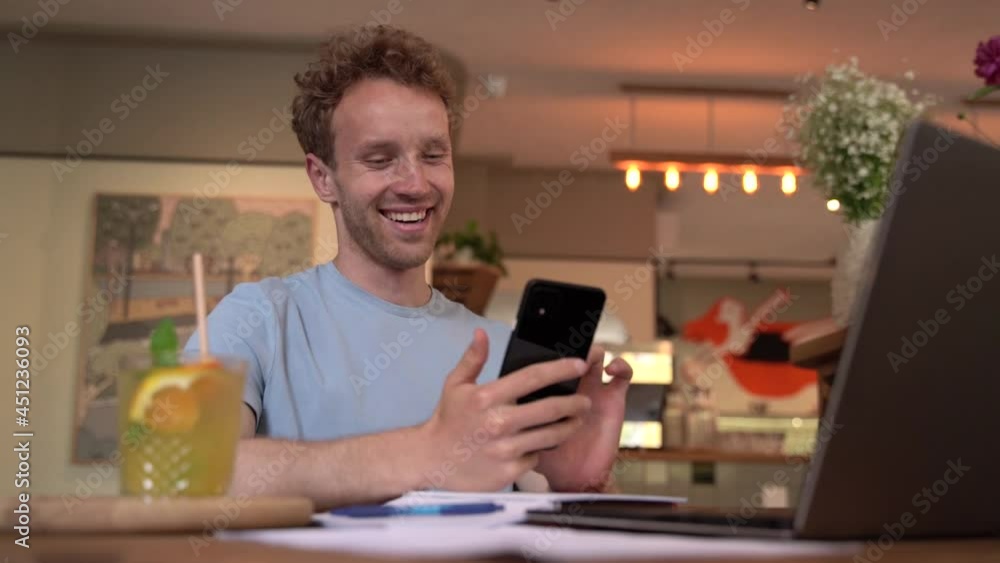 Smiling, positive man sitting at a table in a cafe looking at the phone. Cheerful guy at the table with a laptop Smiling, positive man sitting at a table in a cafe looking at the phone. Cheerful guy at the table with a laptop