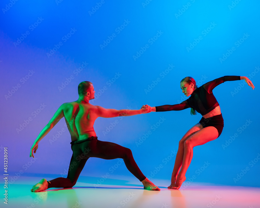 Stylish sportive caucasian couple, male and female dancer dancing contemporary dance on colorful gradient blue pink background in neon light.