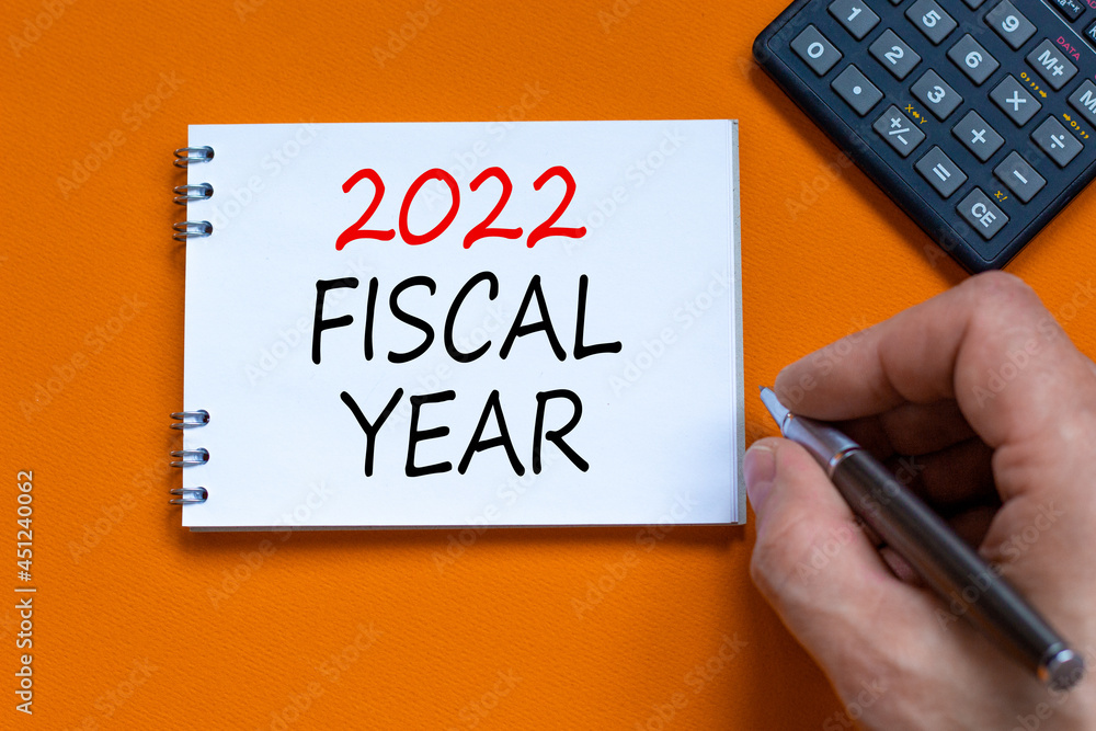 2022 fiscal new year symbol. Businessman writing words '2022 fiscal year' on white note. Black calculator. Beautiful orange background. Business and 2022 fiscal new year concept. Copy space. 2022 fiscal new year symbol. Businessman writing words '2022 fiscal year' on white note. Black calculator. Beautiful orange background. Business and 2022 fiscal new year concept. Copy space.