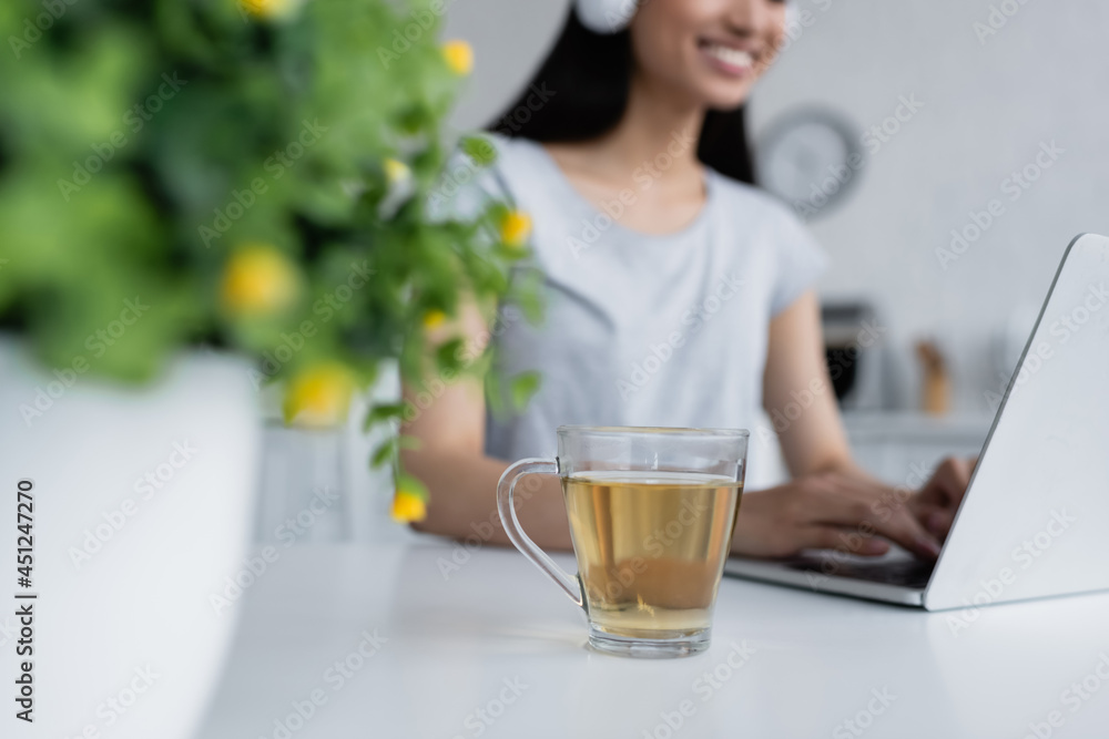 cropped view of blurred asian woman typing on laptop near cup of tea. cropped view of blurred asian woman typing on laptop near cup of tea.