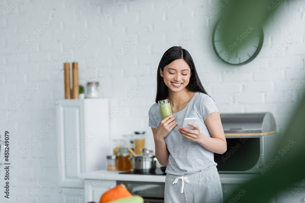 cheerful asian woman with glass of smoothie messaging on cellphone on blurred foreground. cheerful asian woman with glass of smoothie messaging on cellphone on blurred foreground.
