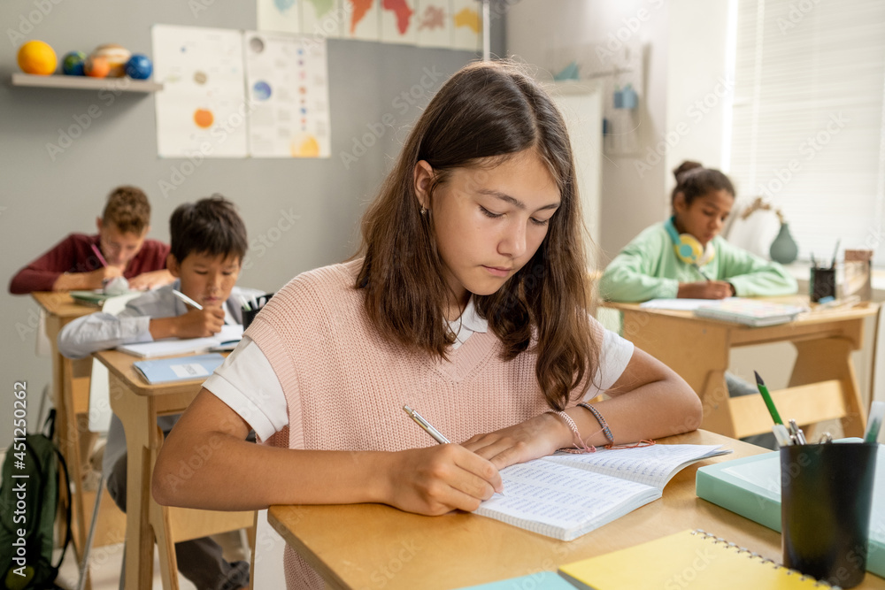 Caucasian elementary schoolgirl making notes while sitting by desk at lesson Caucasian elementary schoolgirl making notes while sitting by desk at lesson