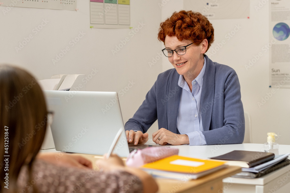 Smiling teacher in eyeglasses using laptop by workplace during lesson Smiling teacher in eyeglasses using laptop by workplace during lesson