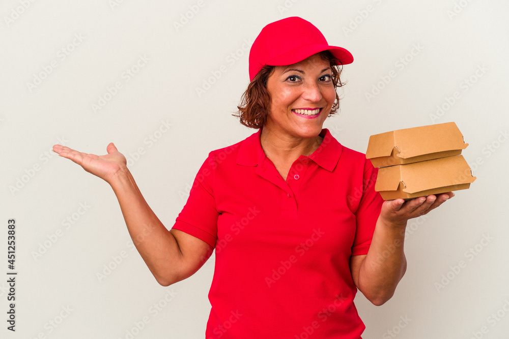 Middle age delivery woman taking burguers isolated on white background showing a copy space on a palm and holding another hand on waist. Middle age delivery woman taking burguers isolated on white background showing a copy space on a palm and holding another hand on waist.