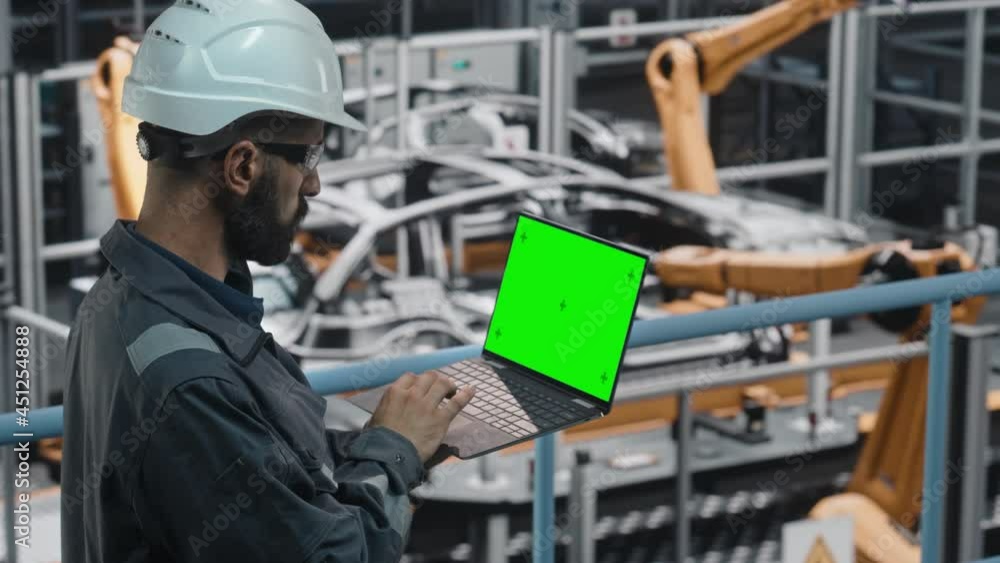 Car Factory Engineer in Work Uniform Using Laptop Computer with Green Screen Mockup Display. Working with Software at Automotive Industrial Manufacturing Facility Dedicated for Vehicle Production. Car Factory Engineer in Work Uniform Using Laptop Computer with Green Screen Mockup Display. Working with Software at Automotive Industrial Manufacturing Facility Dedicated for Vehicle Production.