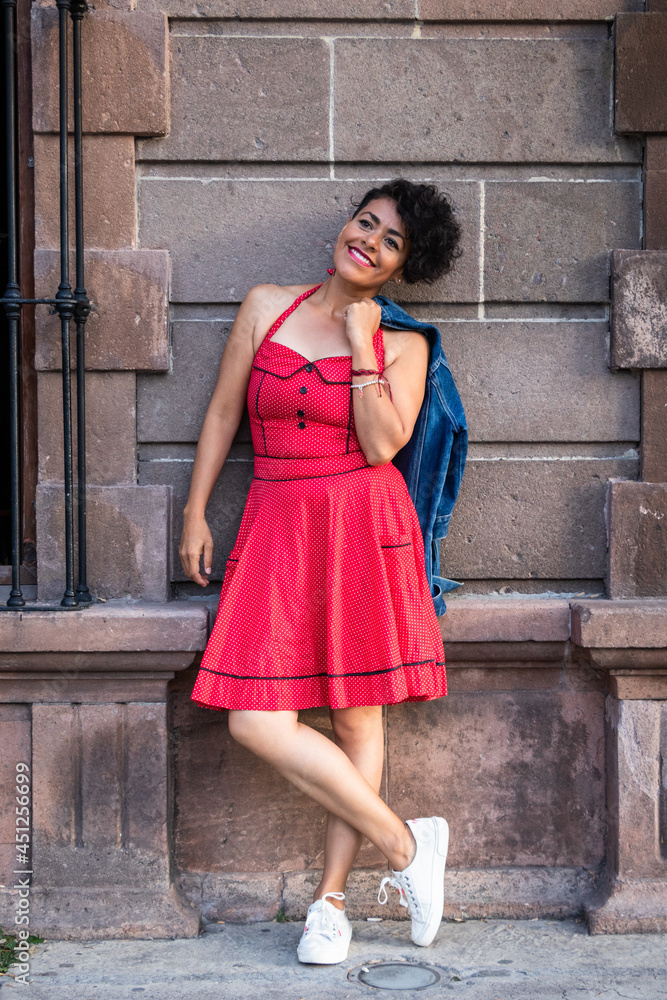 Mid adult afro mexican smiling woman wearing a red dress and white sneakers, leaning against a quarry wall