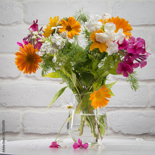 A bouquet of flowers in a glass vase on a white table against a background of a white brick wall.