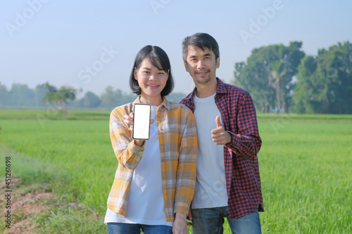 A man and woman, a farmer wearing a striped shirt poses with a smartphone and thumbs up in a field.