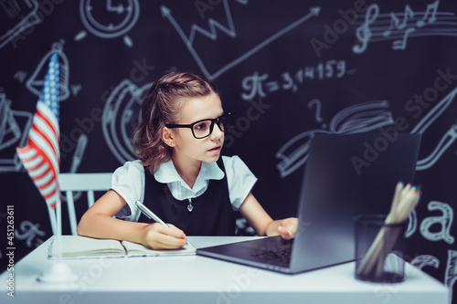beautiful little schoolgirl sitting at desk and study online with laptop against black background with USA flag