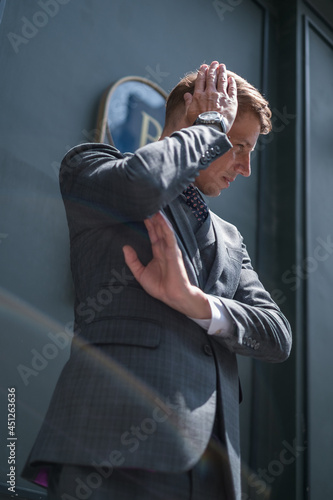 Portrait of handsome young businessman in grey classic suit. Standing near dark green building, touching his head and watching aside. Glare of light