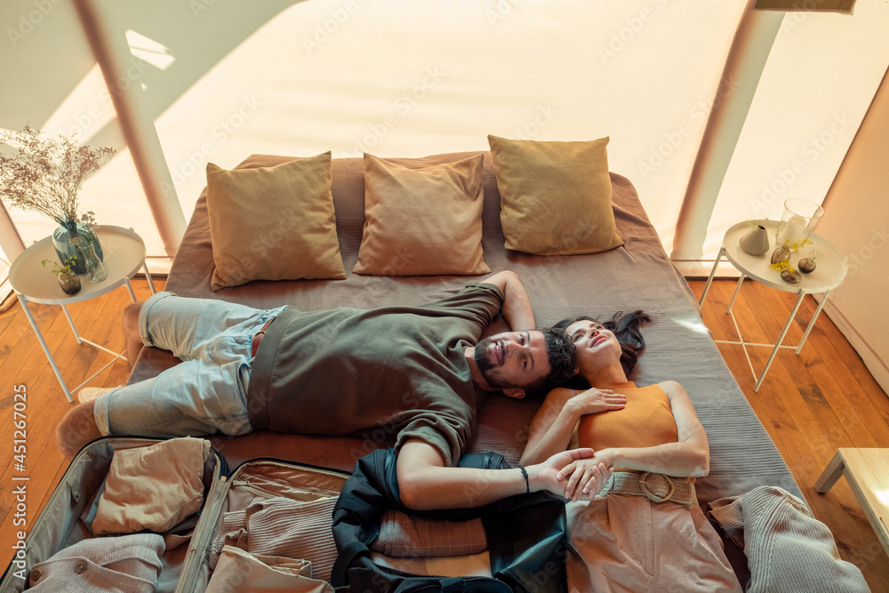 Obraz premium Happy couple daydreaming while lying on bed with unpacked baggage