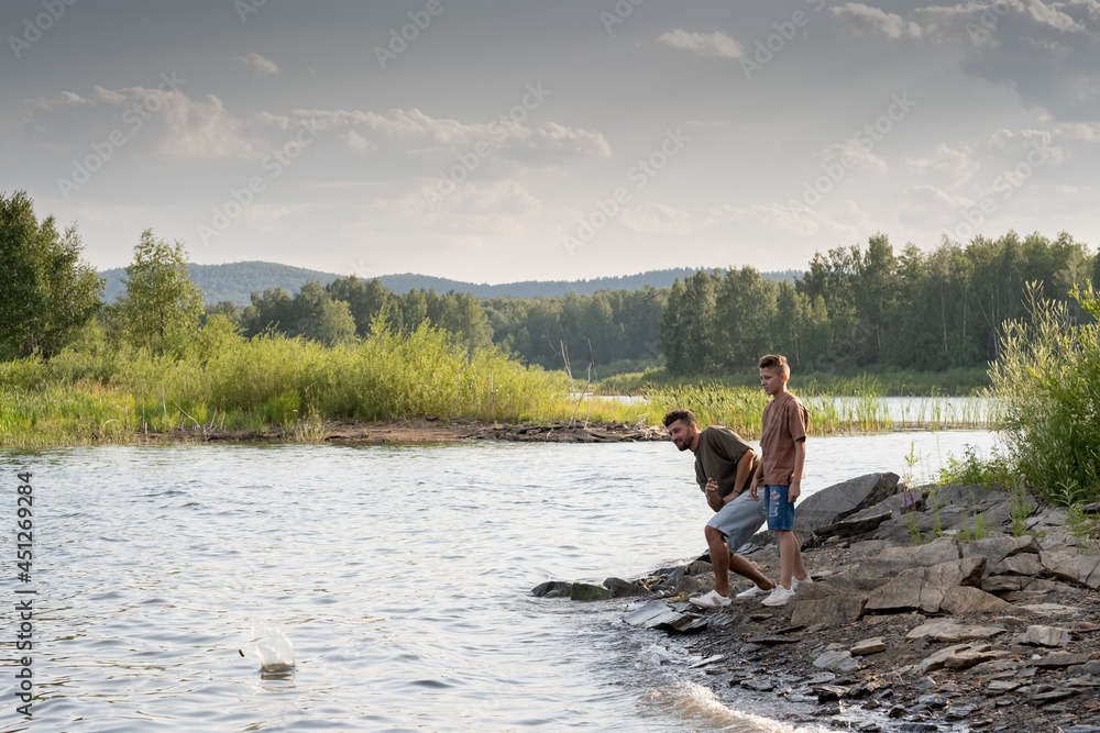 Young man and teenage boy enjoying time by lake and throwing small stones into water