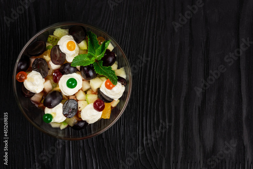 Salad from grapes apples pears kiwi oranges with mascarpone chease and cream. Healthy fresh fruit summer salad in glass bowl on black wooden background with copy space. photo