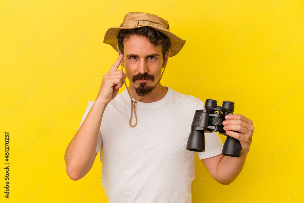 Young caucasian man holding binoculars isolated on yellow background pointing temple with finger, thinking, focused on a task. Young caucasian man holding binoculars isolated on yellow background pointing temple with finger, thinking, focused on a task.