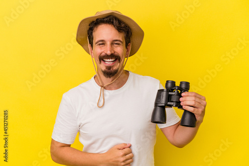 Young caucasian man holding binoculars isolated on yellow background laughing and having fun.