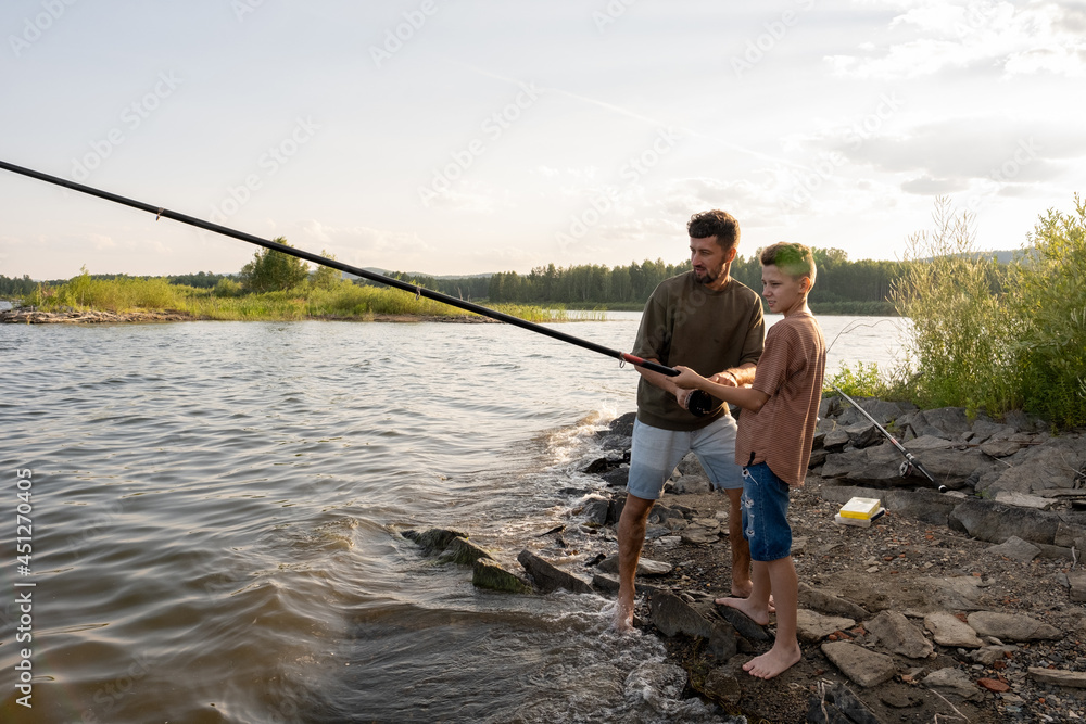 Teenage boy holding long rod while fishing with his father in the countryside Teenage boy holding long rod while fishing with his father in the countryside