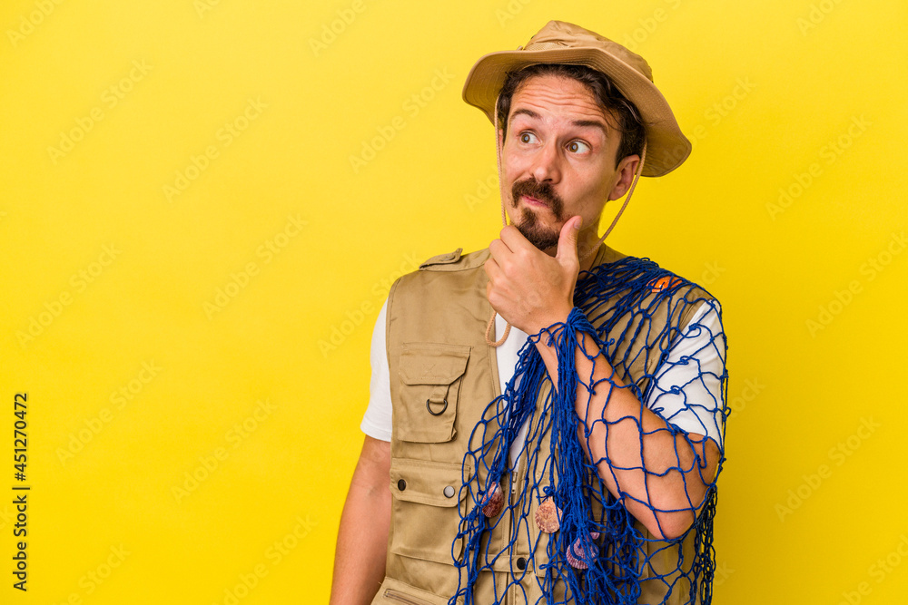 Young caucasian fisherman holding net isolated on yellow background looking sideways with doubtful and skeptical expression.