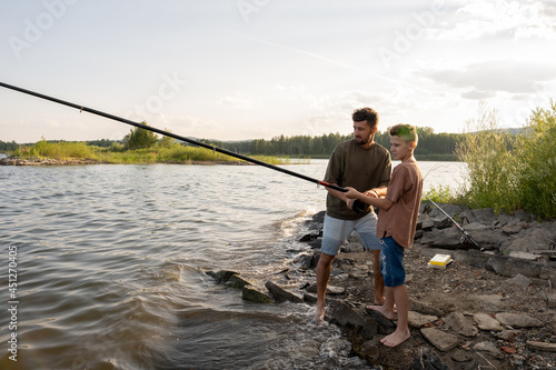 Teenage boy holding long rod while fishing with his father in the countryside