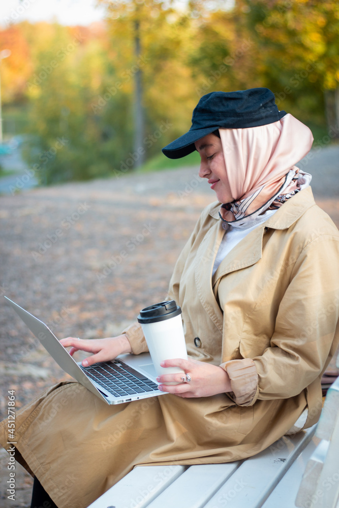 A Muslim woman in a hijab in a beige trench coat sits on a bench with a cup of coffee and a laptop in the park. Muslim business woman. Student. Close plan, face. A Muslim woman in a hijab in a beige trench coat sits on a bench with a cup of coffee and a laptop in the park. Muslim business woman. Student. Close plan, face.
