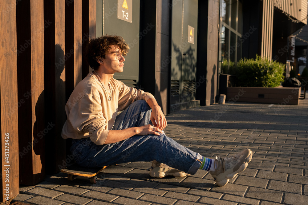 Fototapeta premium Teenage skateboarder sitting by wall on walkside and having rest