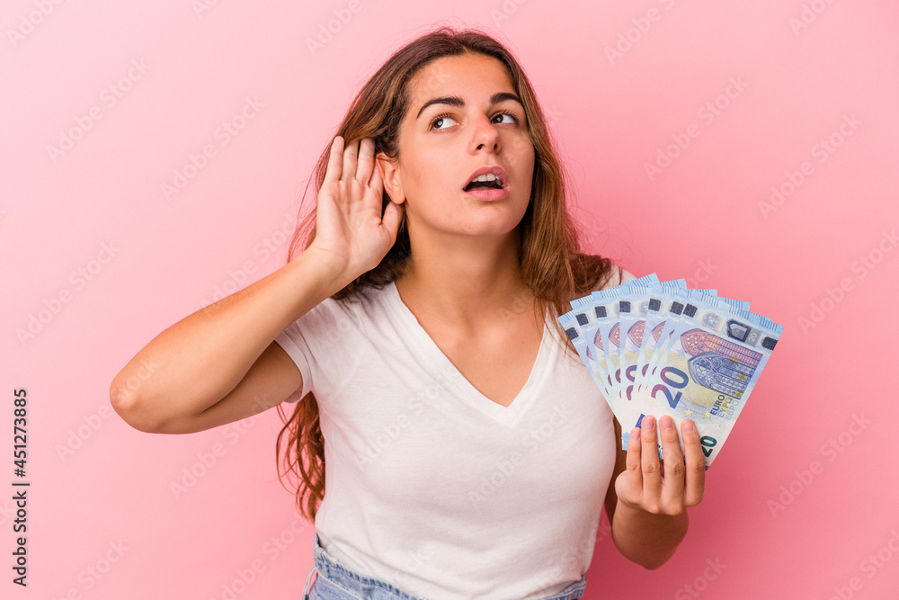 Young caucasian woman holding bills isolated on pink background trying to listening a gossip. Young caucasian woman holding bills isolated on pink background trying to listening a gossip.