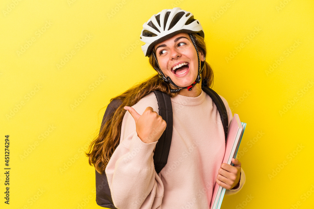 Fototapeta premium Young caucasian student woman wearing a bike helmet isolated on yellow background points with thumb finger away, laughing and carefree.