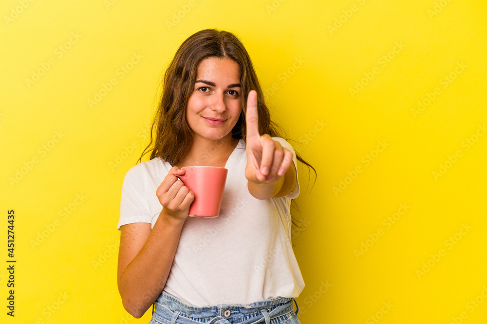 Young caucasian woman holding a mug isolated on yellow background  showing number one with finger.