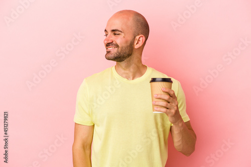 Young caucasian bald man holding a take away coffee isolated on pink background  looks aside smiling, cheerful and pleasant.