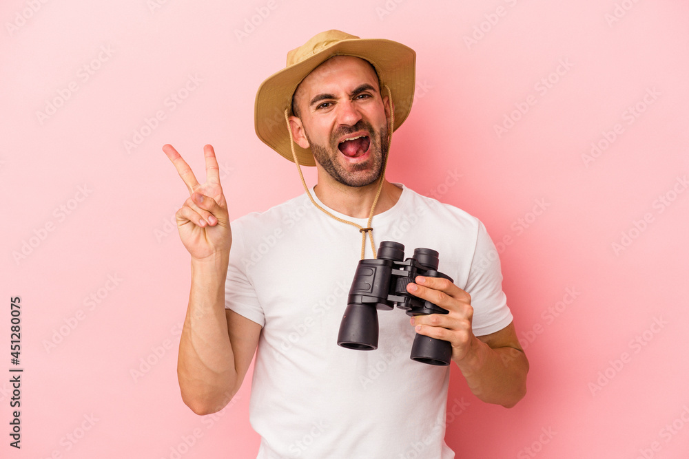 Young caucasian bald man holding binoculars isolated on pink background joyful and carefree showing a peace symbol with fingers. Young caucasian bald man holding binoculars isolated on pink background joyful and carefree showing a peace symbol with fingers.