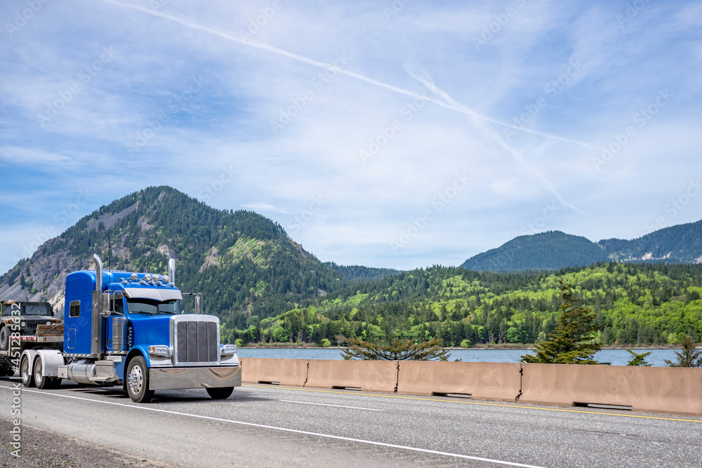 Bright blue big rig classic semi truck tractor with chrome accessories and sleeping cab compartment transporting car on step down semi trailer running on the road along river in Columbia Gorge area