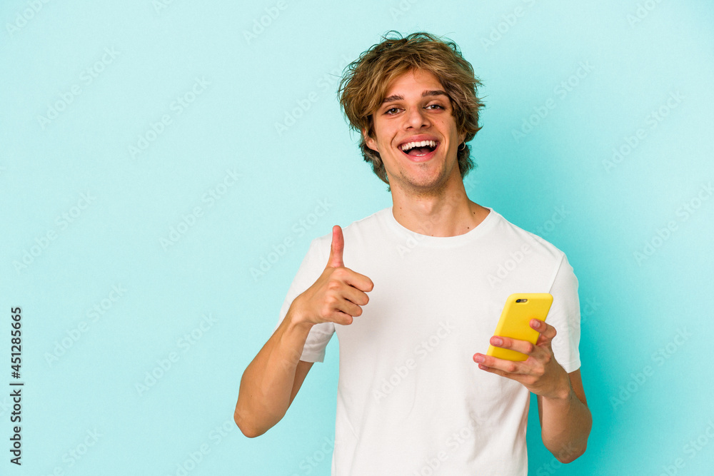 Young caucasian man with makeup holding mobile phone isolated on blue background  smiling and raising thumb up