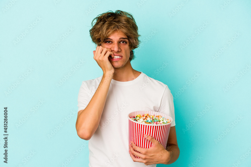 Young caucasian man with makeup holding popcorn isolated on blue background  biting fingernails, nervous and very anxious.