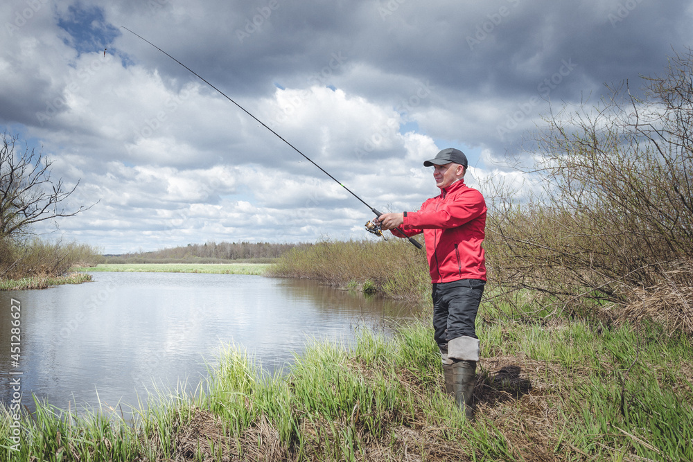 Fisherman in a red jacket is fishing on the green bank of the river. Fisherman in a red jacket is fishing on the green bank of the river.