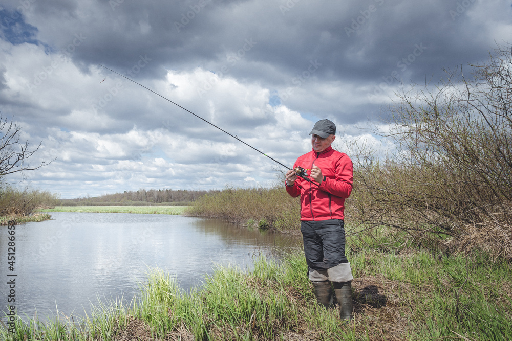 Fisherman in a red jacket is fishing on the green bank of the river.