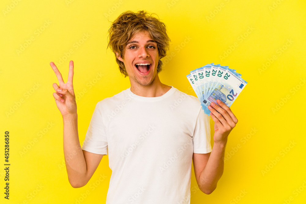 Young caucasian man with makeup holding bills isolated on yellow background showing number two with fingers. Young caucasian man with makeup holding bills isolated on yellow background showing number two with fingers.