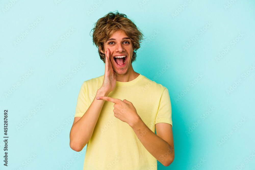 Young caucasian man with make up isolated on blue background  saying a gossip, pointing to side reporting something.